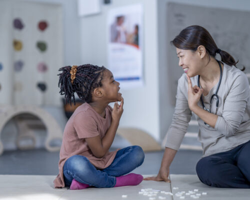 Working On Sounds in Speech Therapy A female therapist sits beside her young patient on the floor as they work on some speech therapy exercises. They are each dressed casually and have their hands on their chins as they work through phonetical sounds. Small white letter tiles can be seen laying out in front of them from a previous exercise.